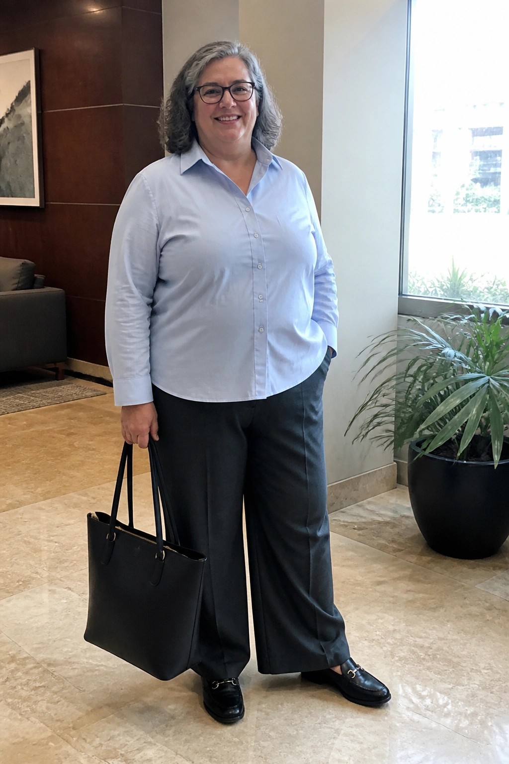 Woman with gray hair in blue shirt and black pants holding black tote bag indoors