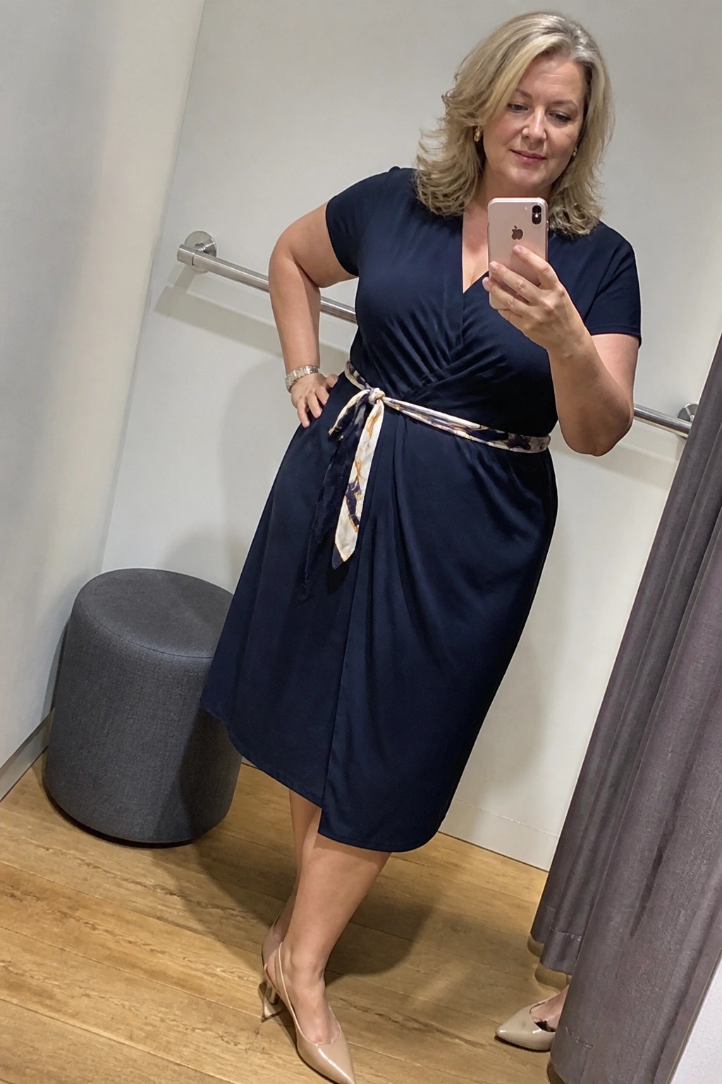 Curvy blonde woman in navy wrap dress taking mirror selfie with phone