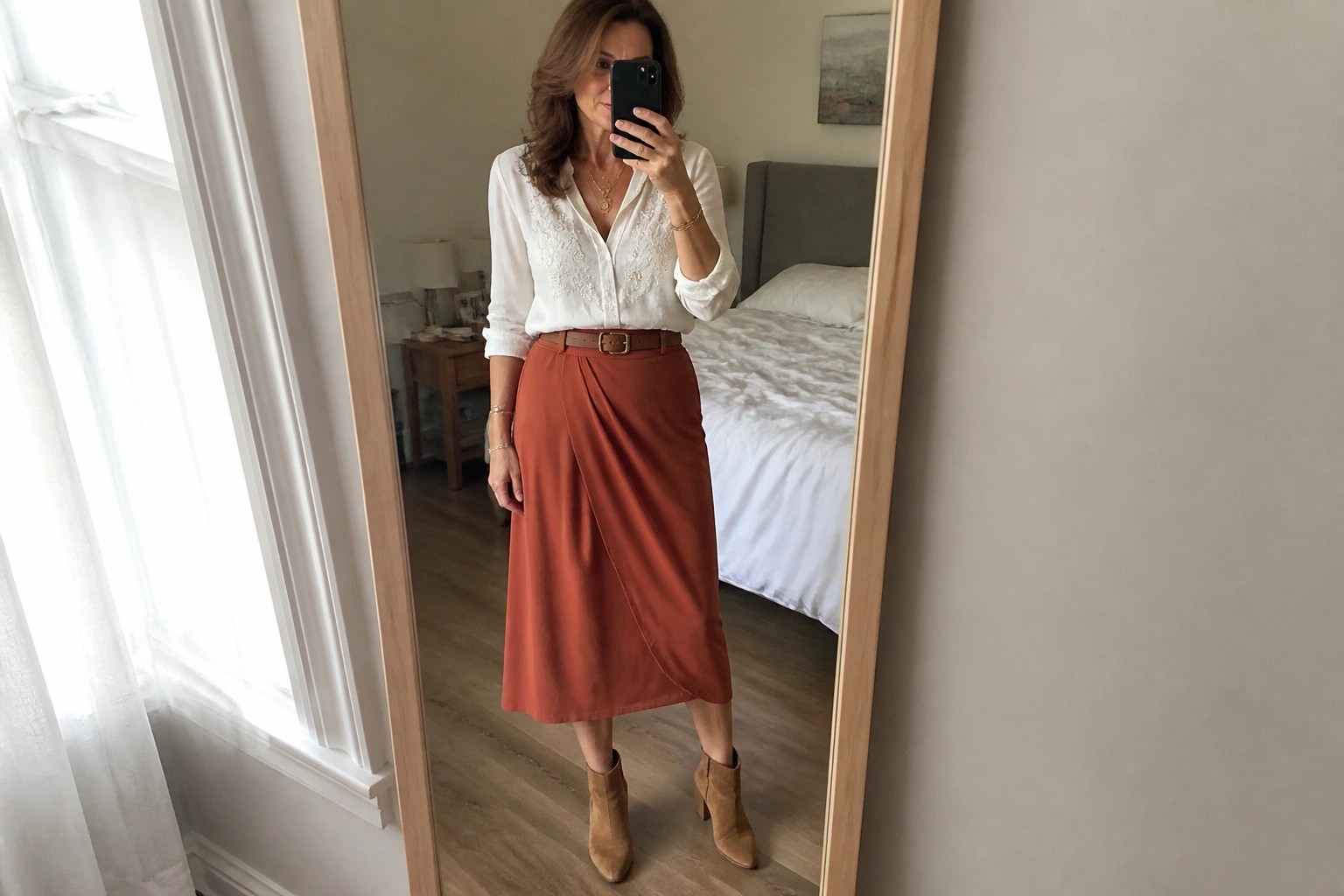 Woman in white blouse and rust midi skirt taking bedroom mirror selfie
