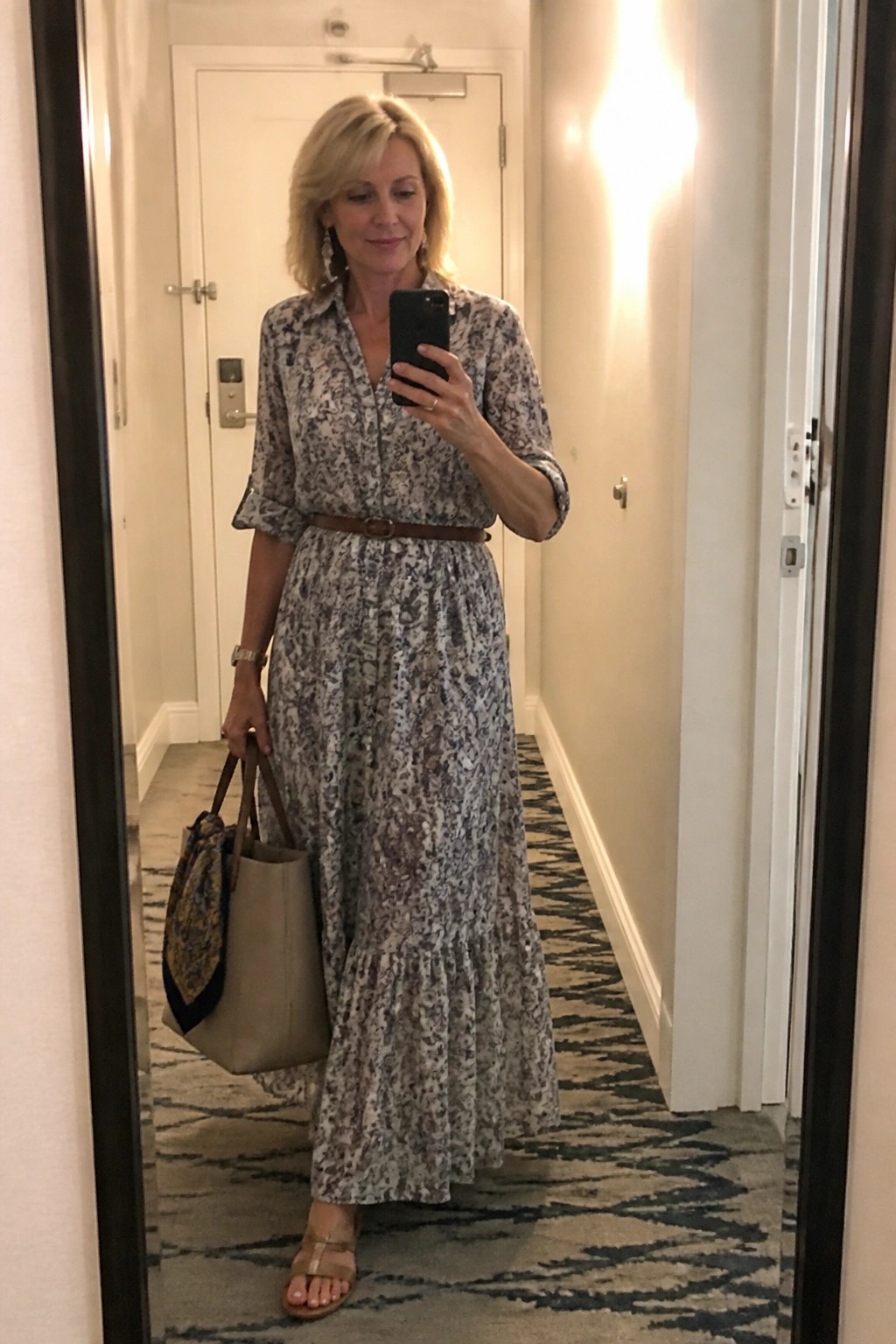 Blonde woman in belted patterned maxi dress takes mirror selfie holding tote bag