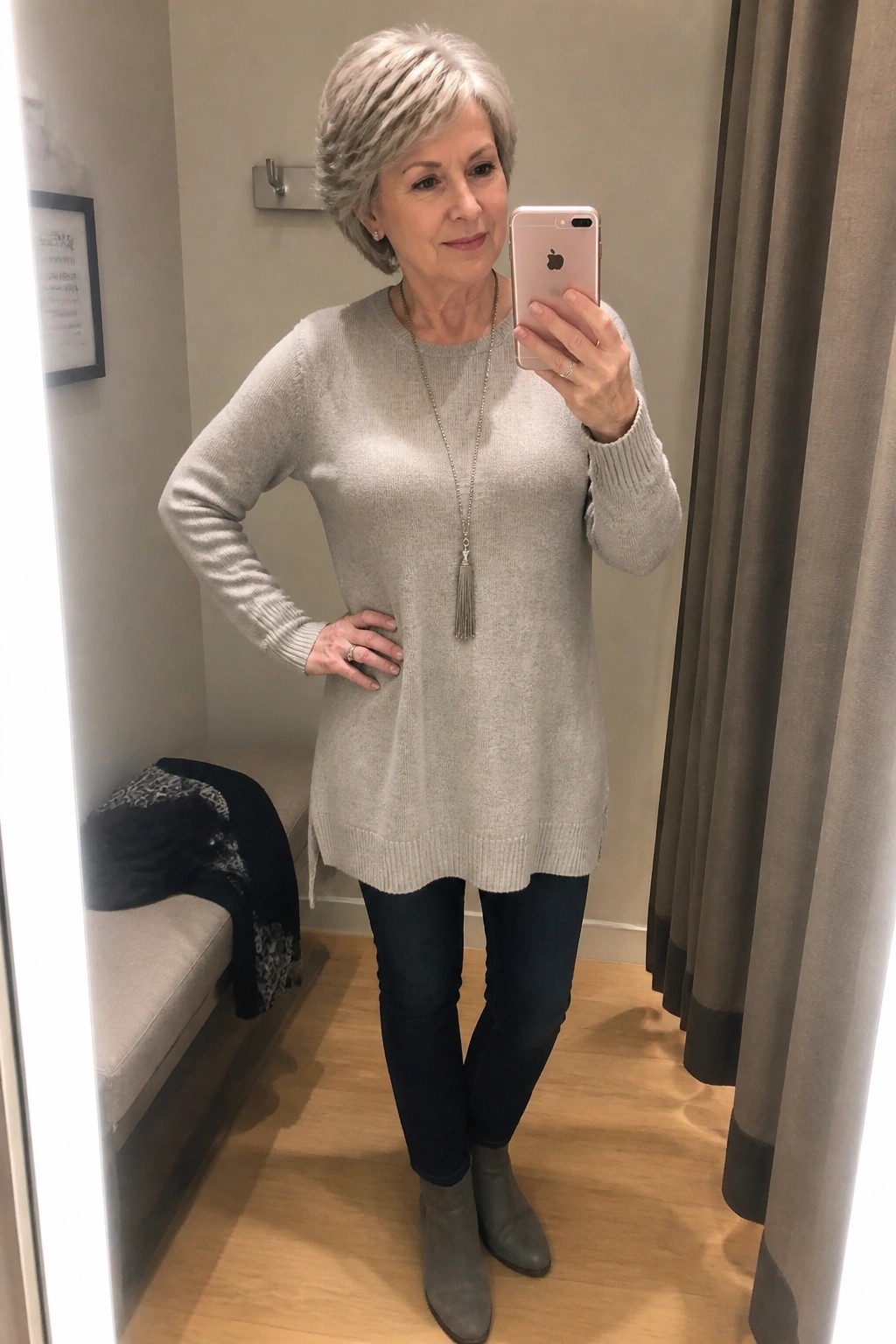 Silver-haired woman in gray sweater taking mirror selfie in fitting room with jeans and boots