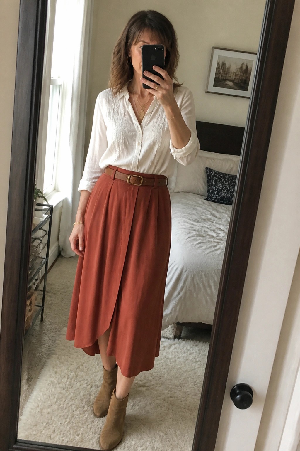 Woman in white blouse and rust midi skirt taking bedroom mirror selfie