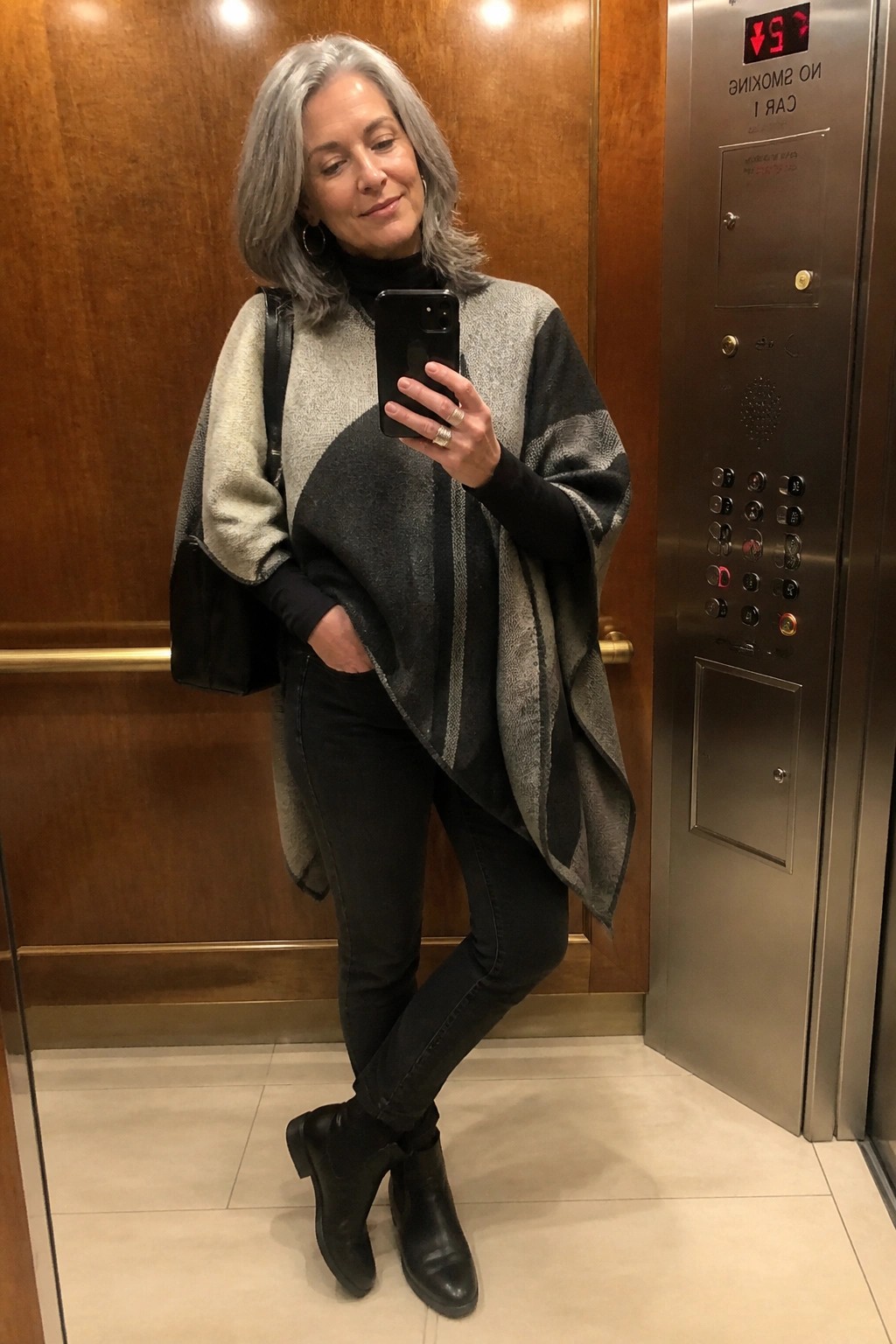 Woman over 50 in elevator mirror selfie wearing gray and black patterned poncho over black turtleneck, black jeans, and black ankle boots with a black shoulder bag.