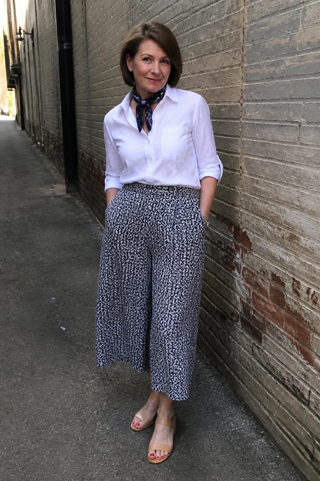 Woman in white shirt, patterned scarf and pants stands in brick alleyway.
