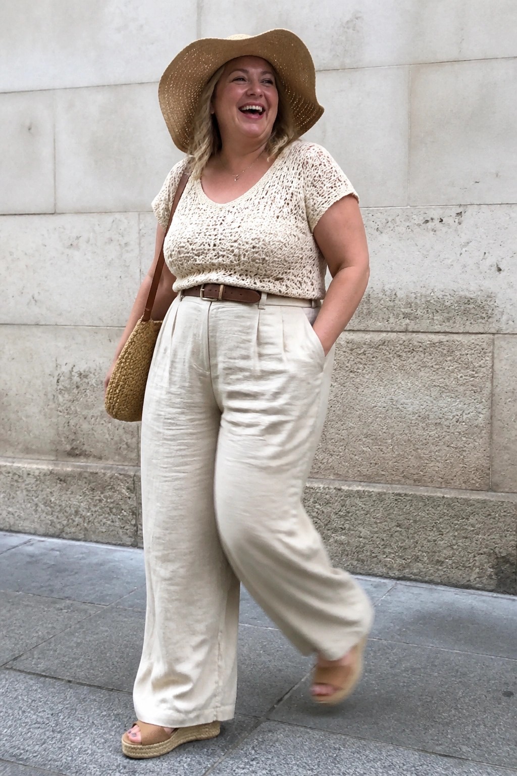 Woman in cream crochet top, wide leg linen pants, brown belt, straw hat, woven bag, and espadrille sandals standing against a stone wall