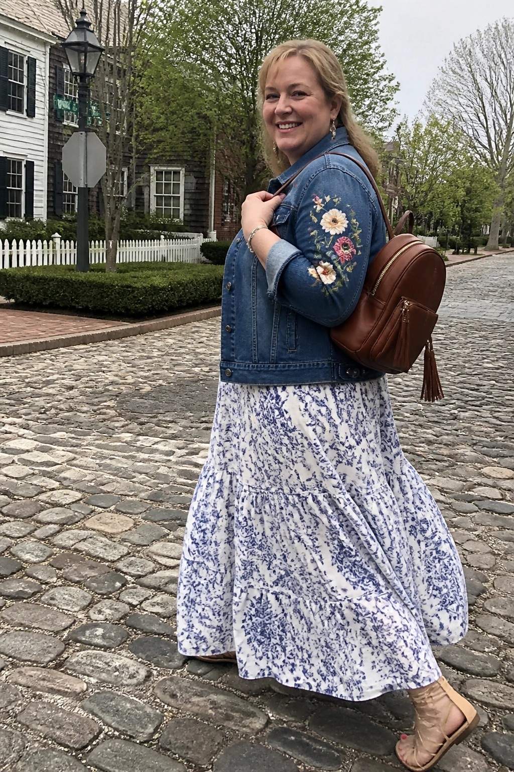 Woman in blue embroidered denim jacket and white-blue floral tiered dress on cobblestones