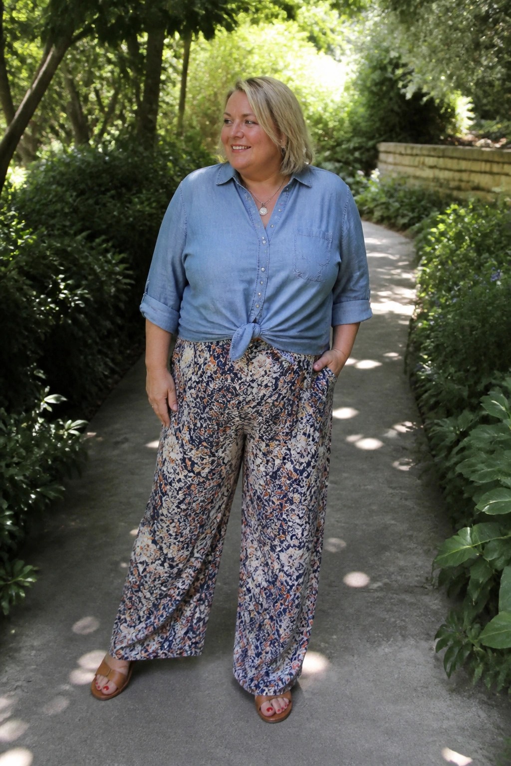 Blonde woman in knotted blue shirt and floral wide-leg pants on garden path.