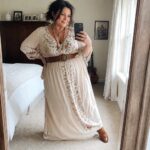 Woman in cream floral maxi dress with brown belt and boots taking bedroom mirror selfie