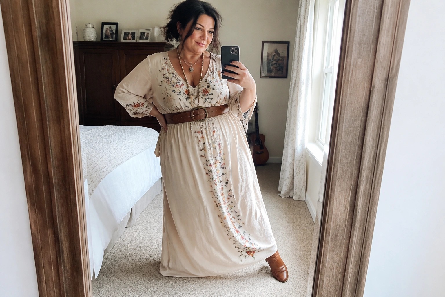 Woman in cream floral maxi dress with brown belt and boots taking bedroom mirror selfie