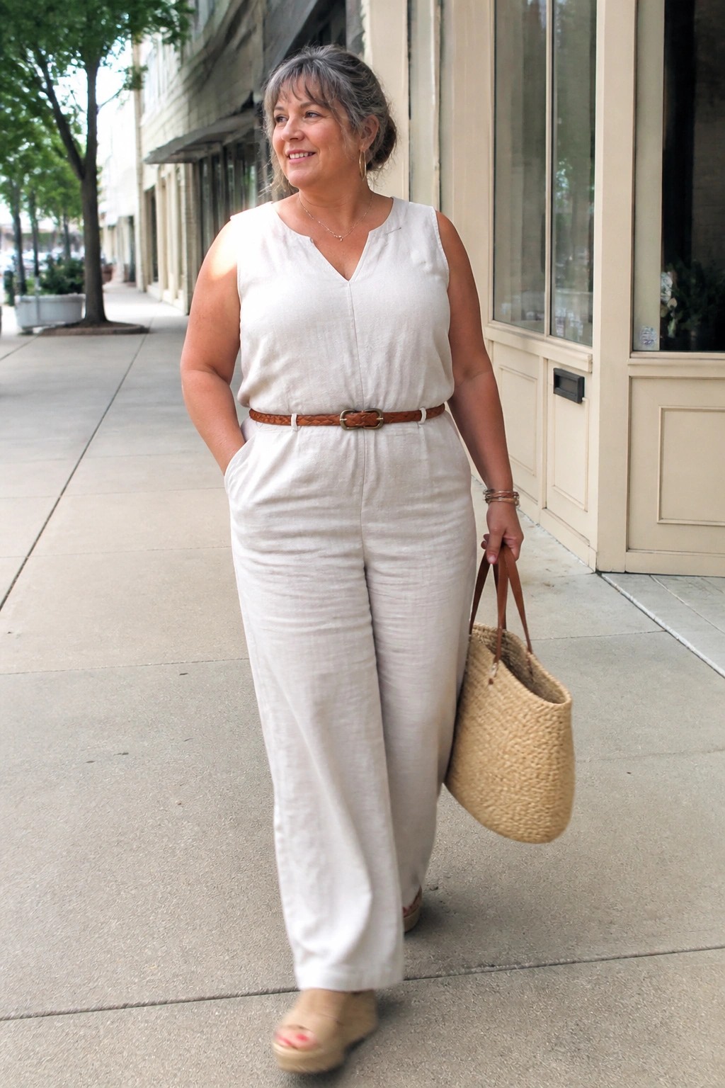 Woman in white jumpsuit with brown belt holding woven straw bag on sidewalk