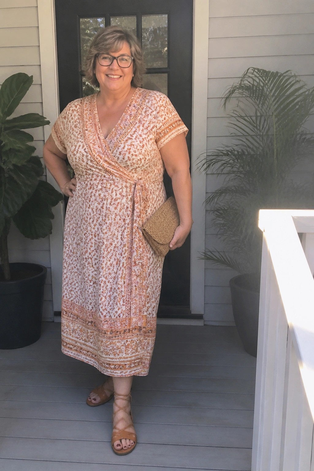 Woman in white-orange patterned wrap dress holding woven clutch on porch