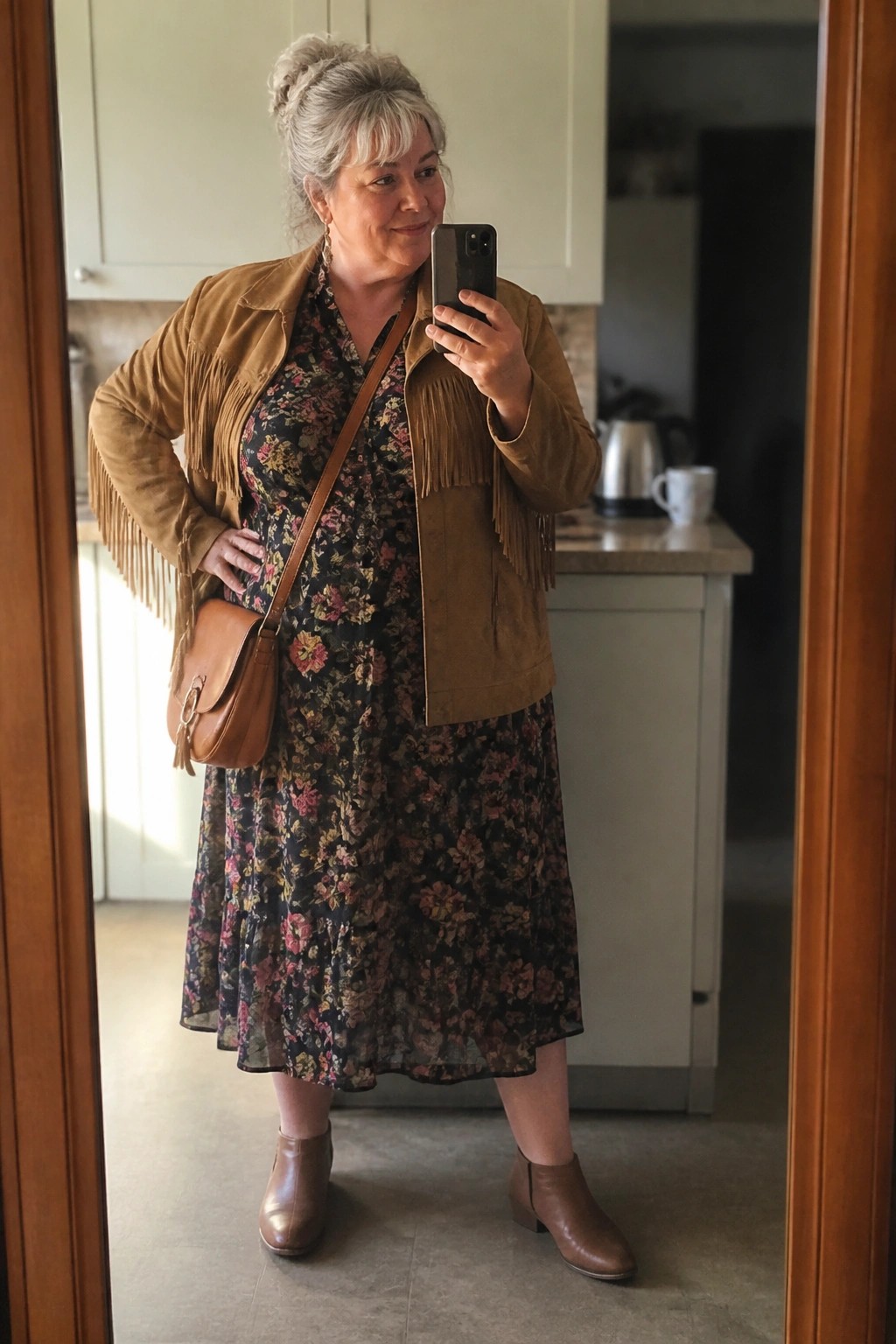 Woman in floral dress and fringed suede jacket taking mirror selfie in kitchen