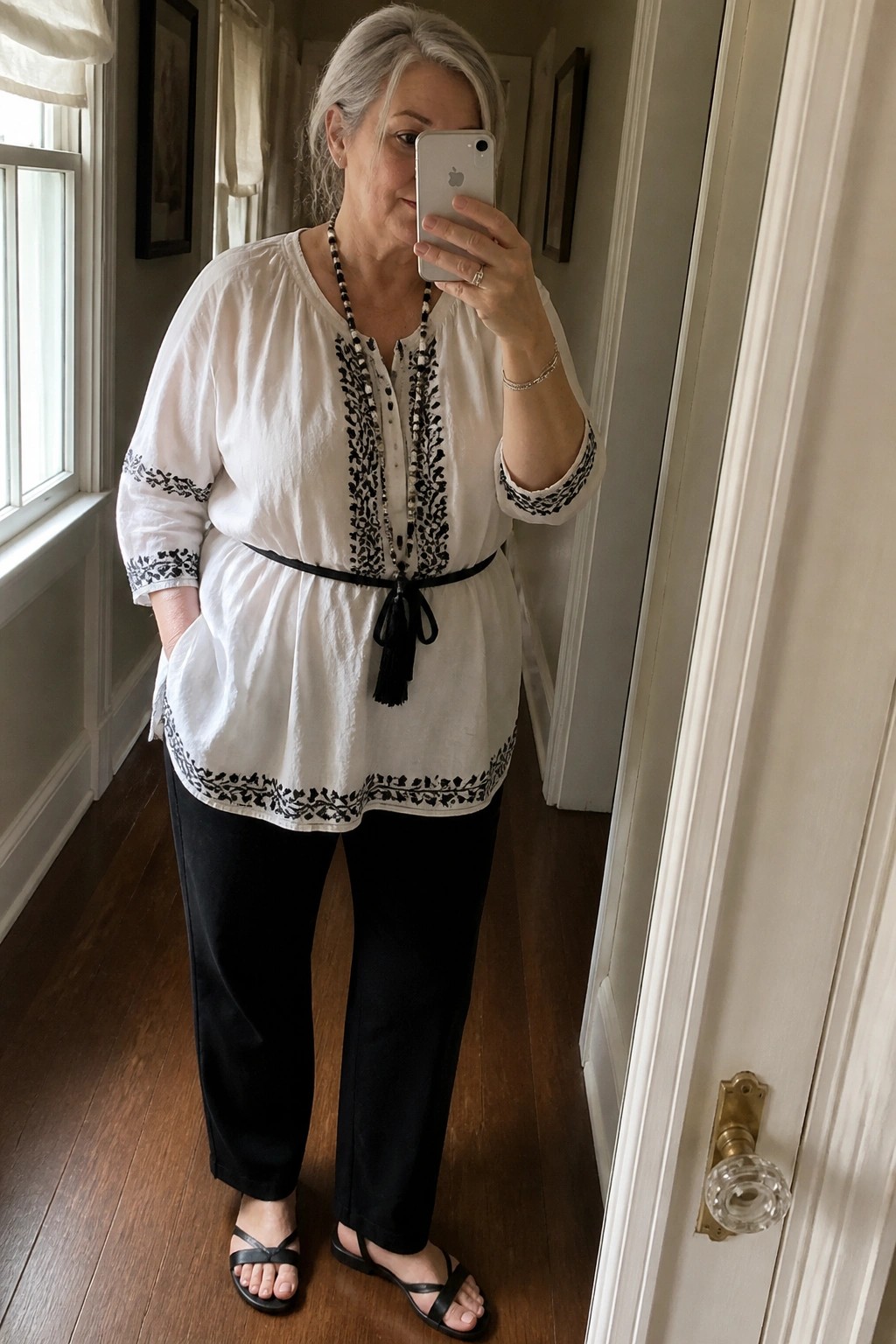 Gray-haired woman in white embroidered blouse with black belt takes mirror selfie.
