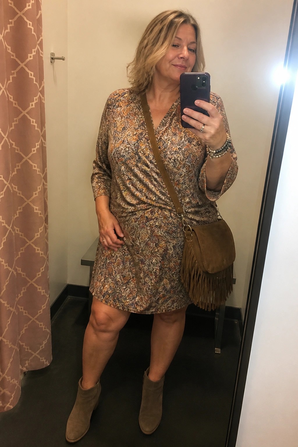 Blonde woman in patterned wrap dress with fringed suede bag taking mirror selfie in fitting room