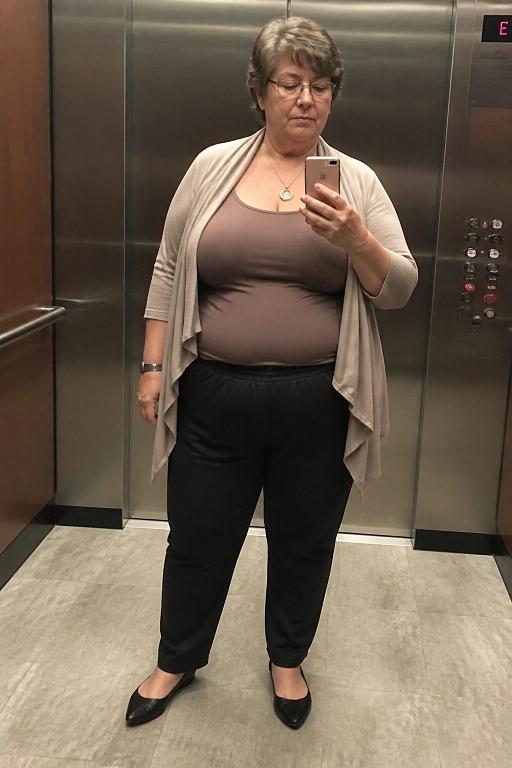 Woman in beige cardigan holding smartphone in elevator wearing black pants and flats