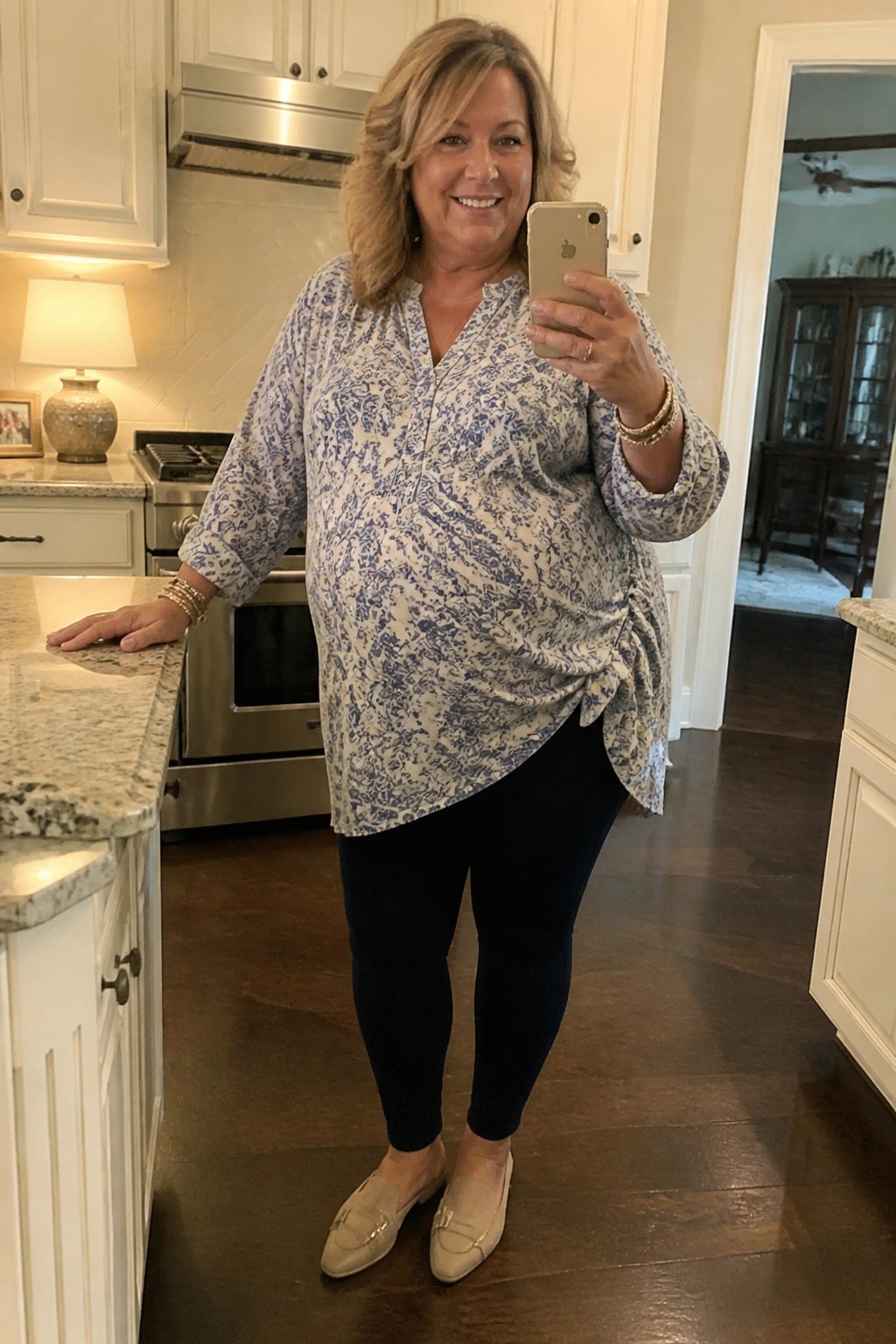 Smiling blonde woman in blue patterned top taking selfie in kitchen.