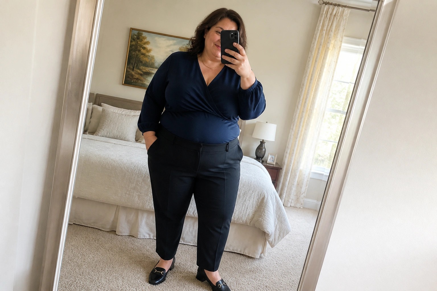 Woman in navy wrap top and black pants taking bedroom mirror selfie with phone