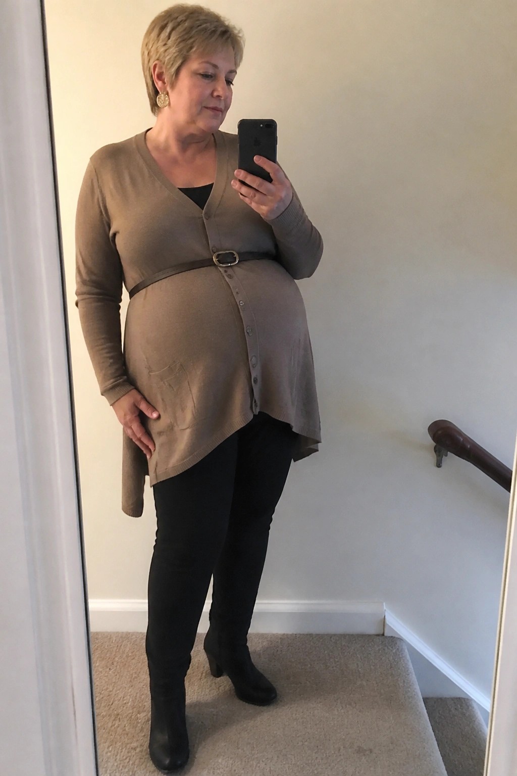 Pregnant woman with short blonde hair takes mirror selfie in beige cardigan and black boots