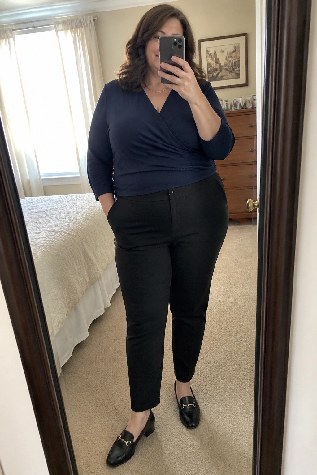 Woman in navy wrap top and black pants taking bedroom mirror selfie with phone