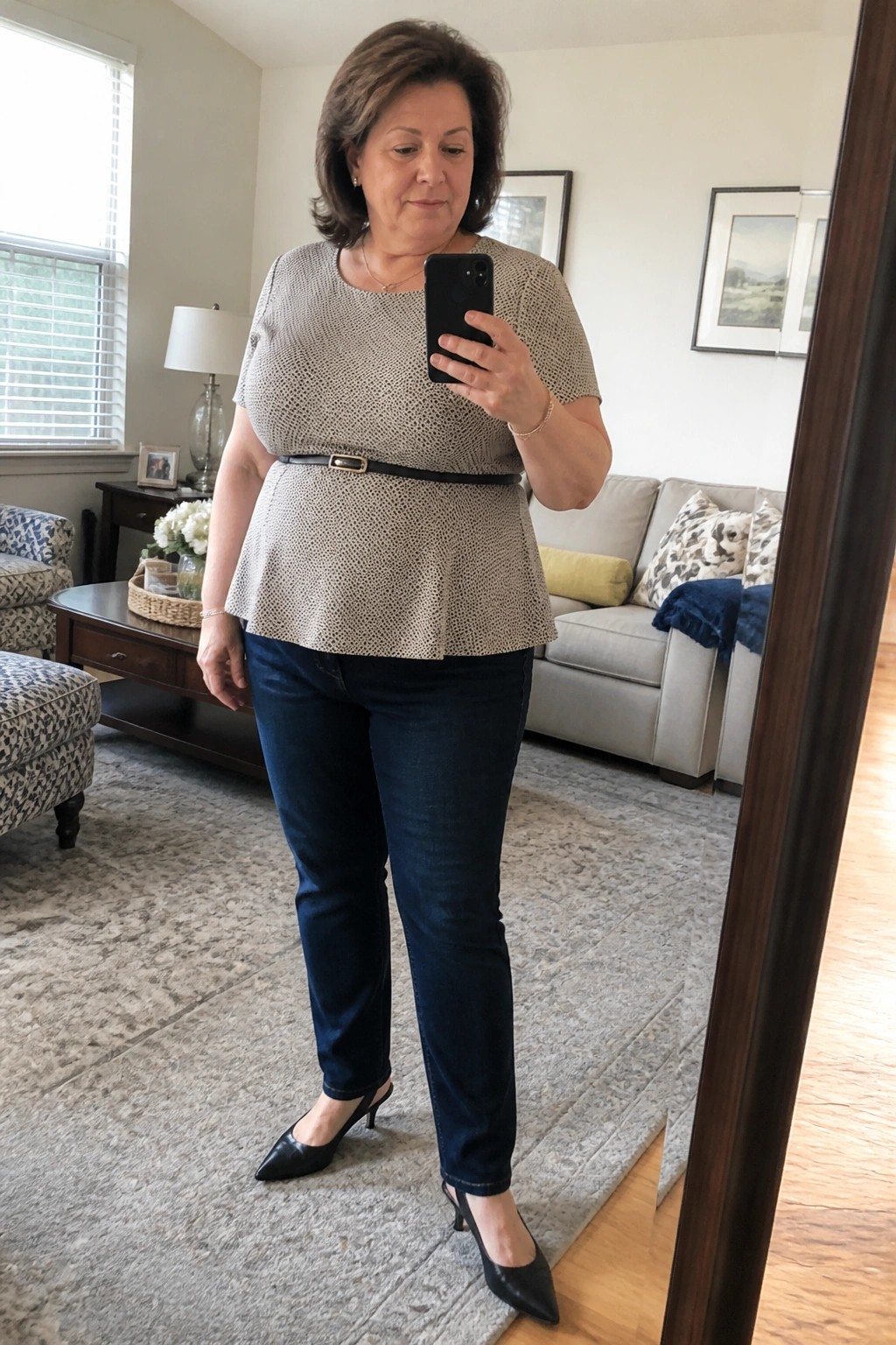 Woman in dotted beige peplum top with black belt, dark jeans, and black kitten heels standing in a living room