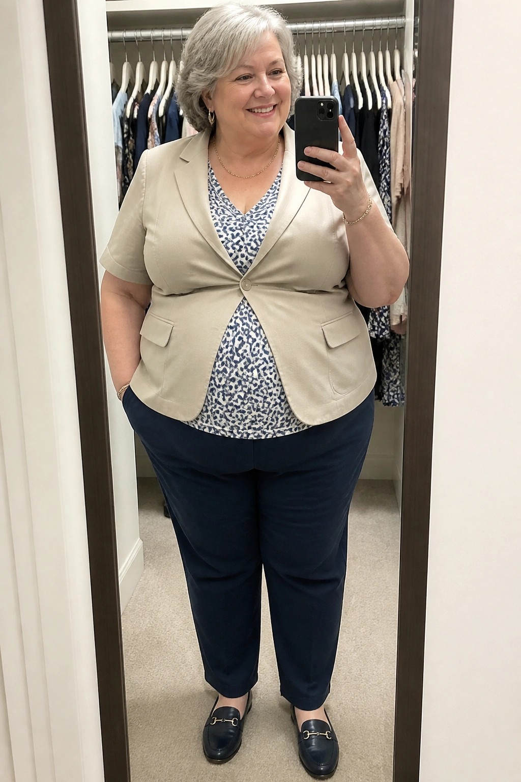 Smiling gray-haired woman in beige blazer and navy pants taking mirror selfie in closet.