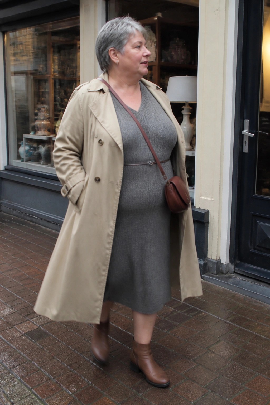 Woman with short gray hair in beige trench coat over gray knit dress on brick sidewalk