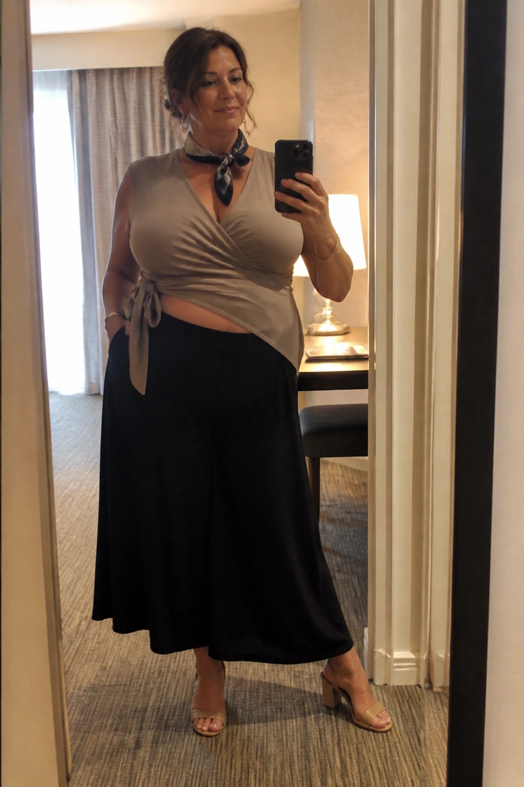 Woman in taupe wrap top and black wide-leg pants taking mirror selfie in hotel room