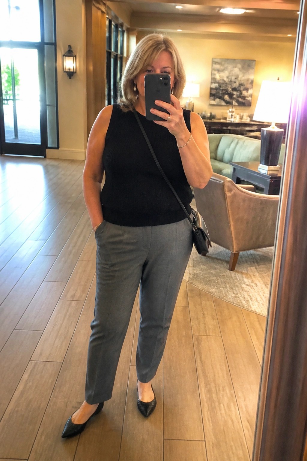 A woman over 50 wearing a black sleeveless knit tank top with gray tailored pants and black pointed flats while taking a mirror selfie in a lobby.