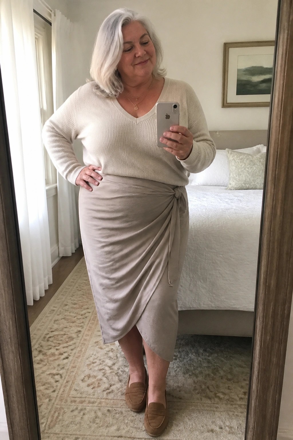Plus size woman with white hair in a cream V neck sweater paired with a taupe wrap midi skirt and brown suede loafers taking a mirror selfie indoors.