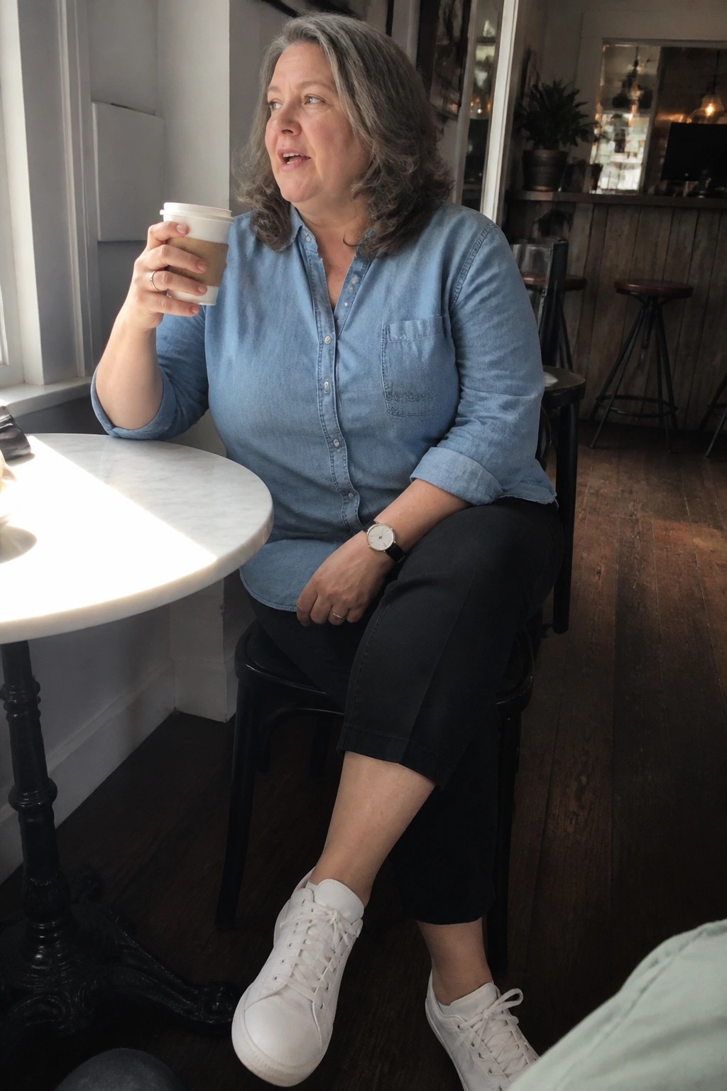 Plus size woman over 50 in light blue denim button up shirt black cropped pants and white sneakers holding a coffee cup while seated indoors.