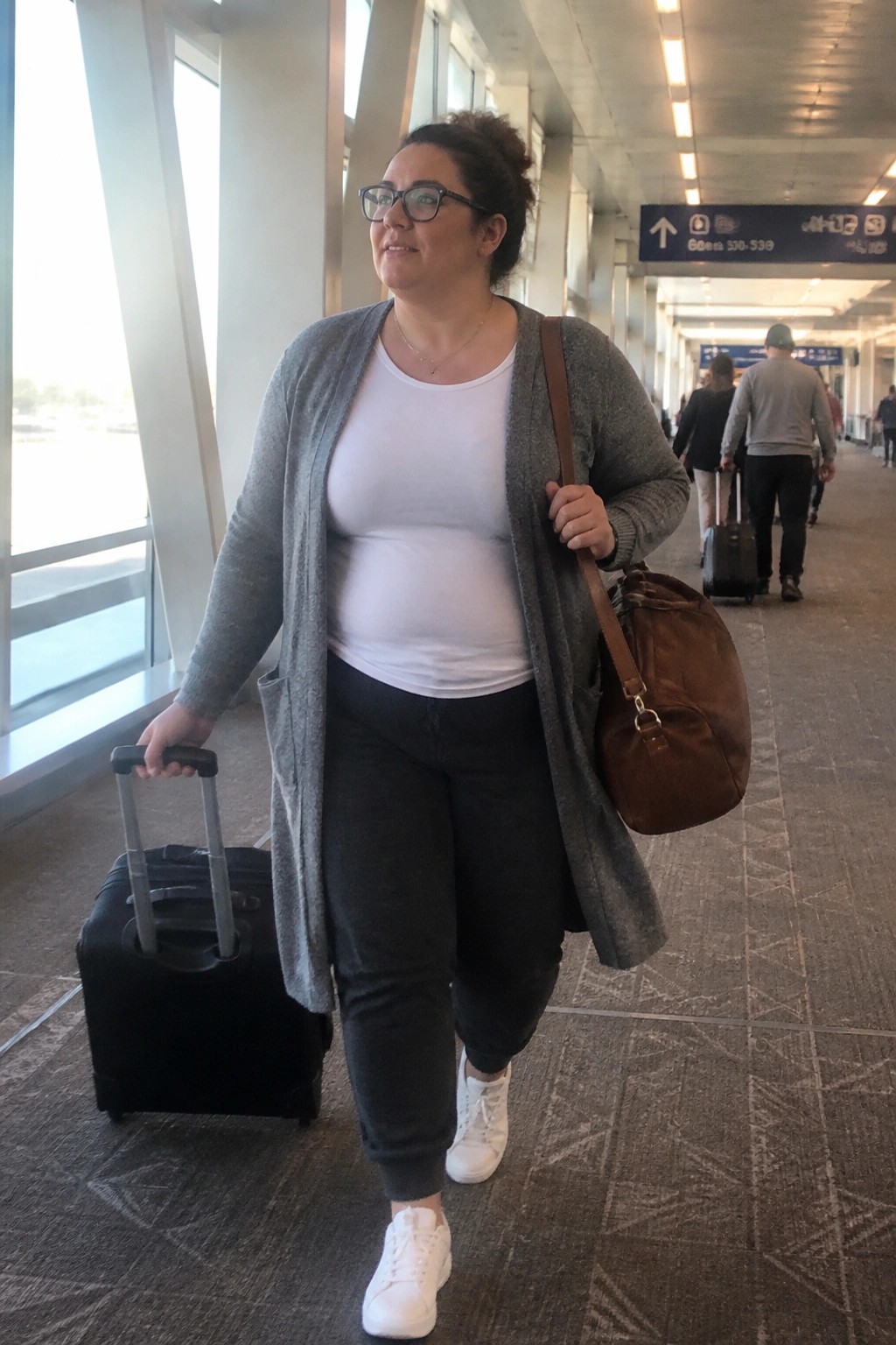 Plus size woman in gray open cardigan over white tee and black pants with white sneakers pulling a black rolling suitcase and carrying a brown bag in an airport hallway.