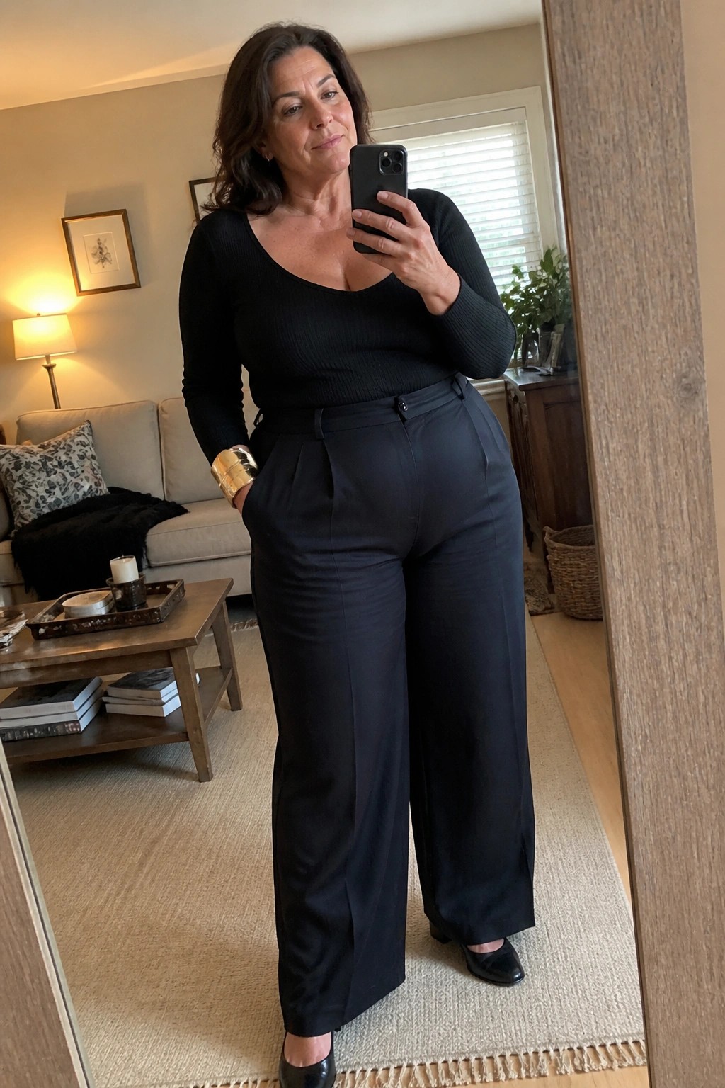 Woman in black ribbed top and wide-leg pants taking mirror selfie in living room