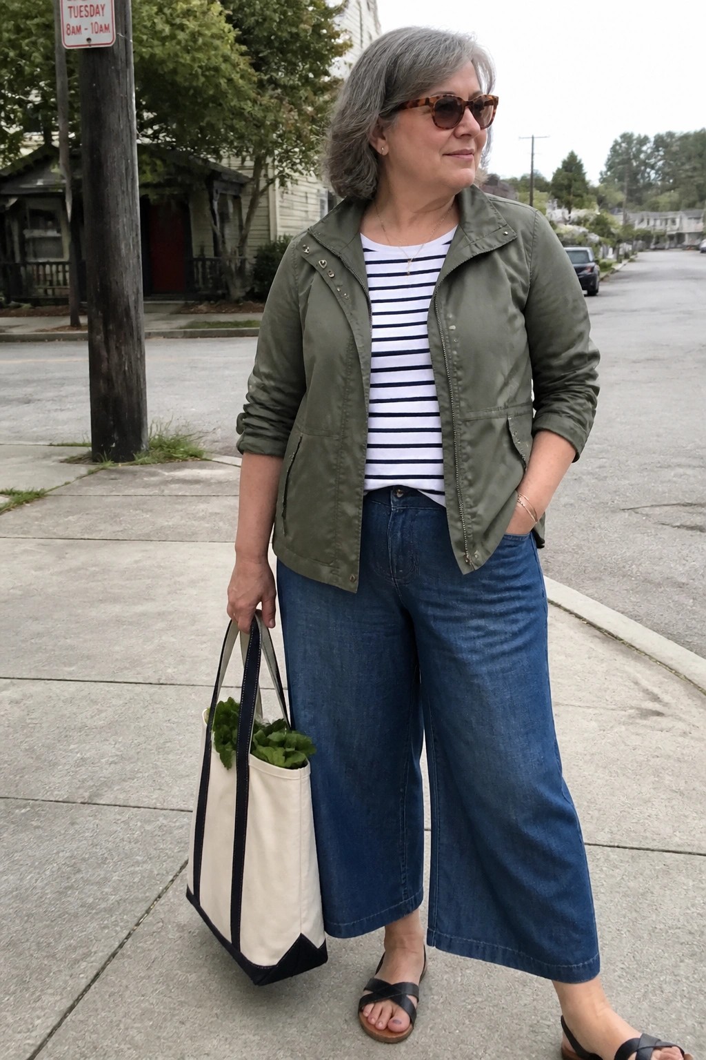 Woman over 50 wearing an olive utility jacket over a navy and white striped tee with blue wide leg denim culottes, black strappy sandals, and carrying a canvas tote bag.