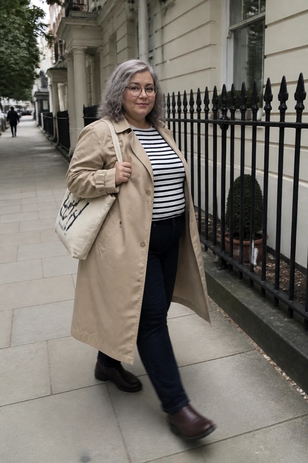 A plus size woman with gray hair wearing a beige trench coat over a black and white striped shirt, dark jeans, and brown boots while carrying a tote bag on a city sidewalk.