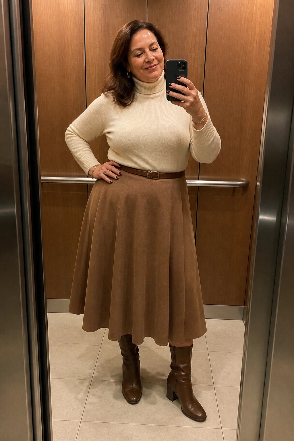 Woman in cream turtleneck, brown skirt and boots taking selfie in elevator.