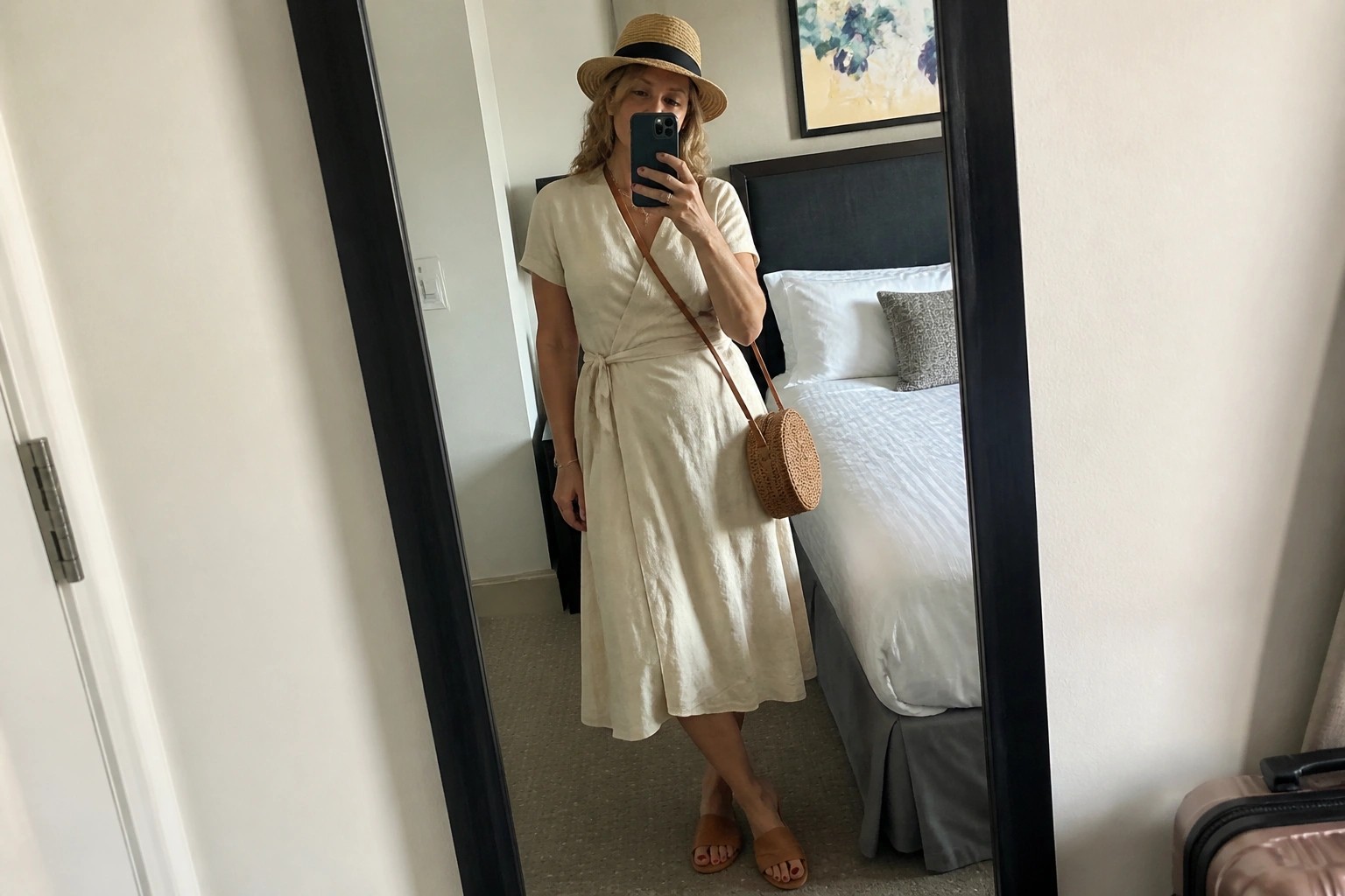 Woman in beige wrap dress and straw hat taking mirror selfie in hotel room