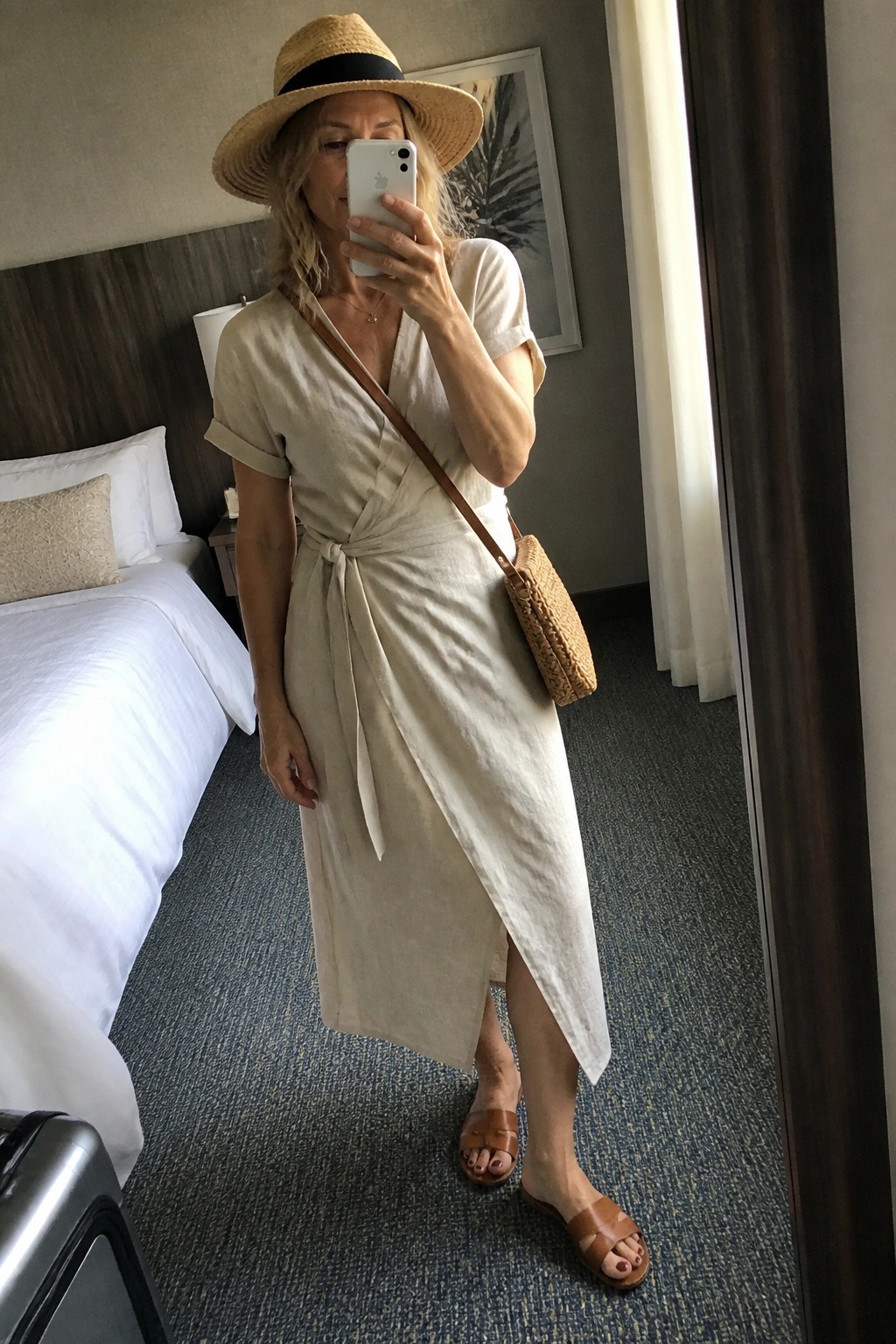 Woman in beige wrap dress and straw hat taking mirror selfie in hotel room