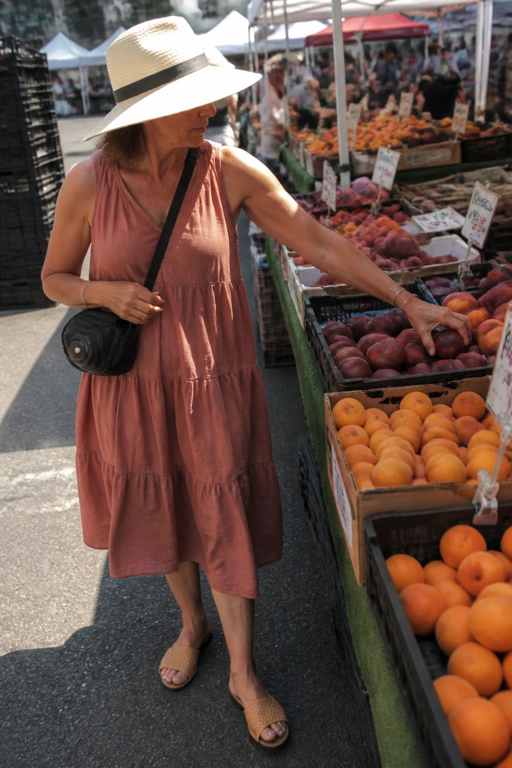 Woman over 50 wearing a dusty rose tiered sleeveless midi dress with a black crossbody bag, straw hat, and beige woven slide sandals while reaching for fruit at an outdoor market.