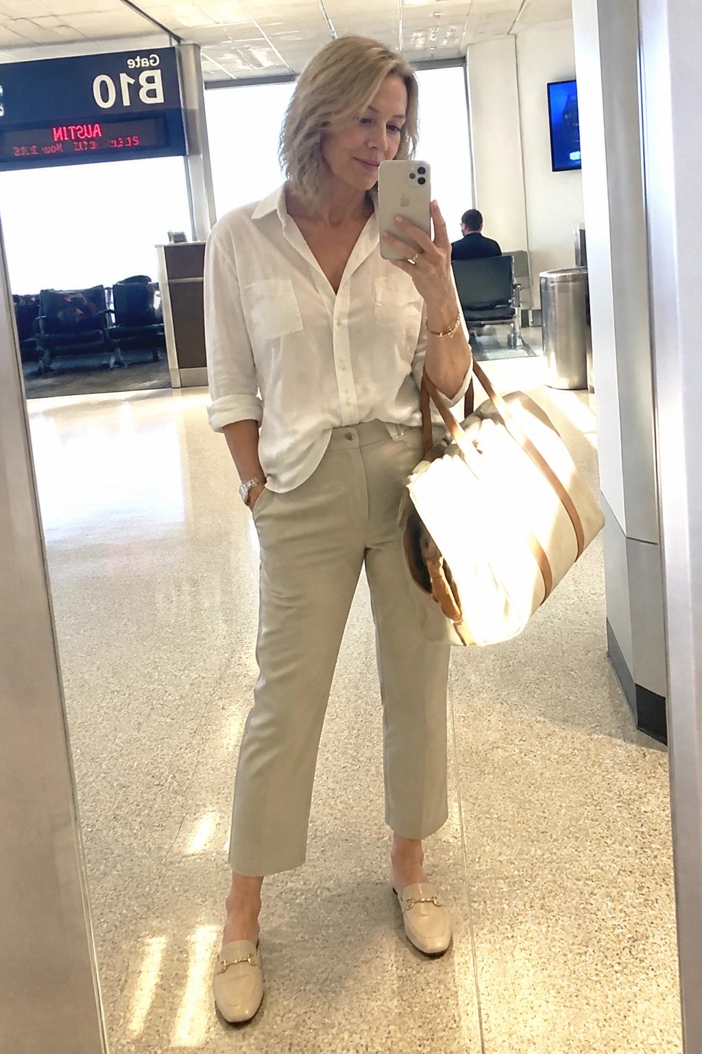 Woman in white shirt and beige pants taking mirror selfie at airport gate with tote bag