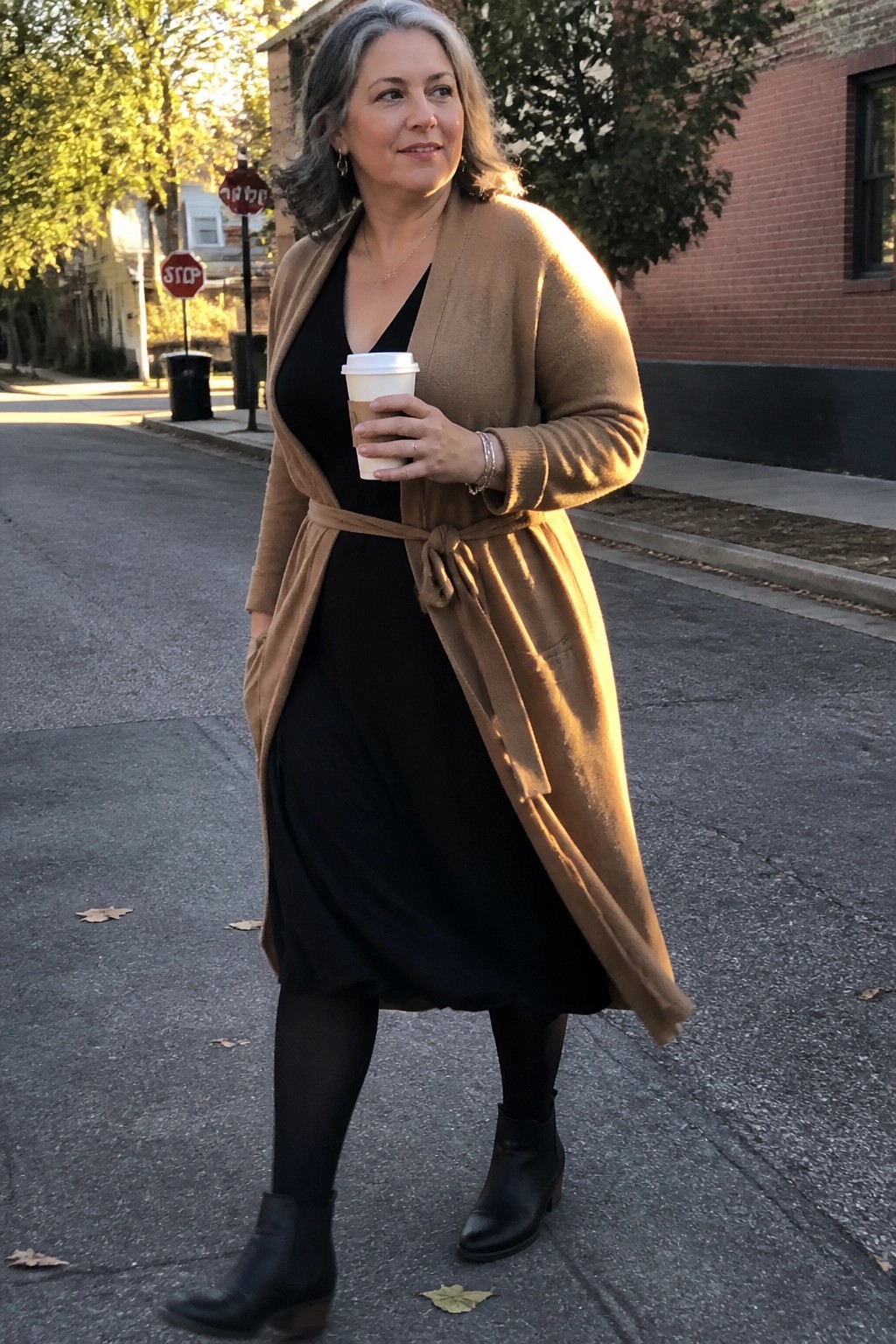Woman wearing a long tan belted cardigan over a black midi dress with black tights and ankle boots while holding a coffee cup.