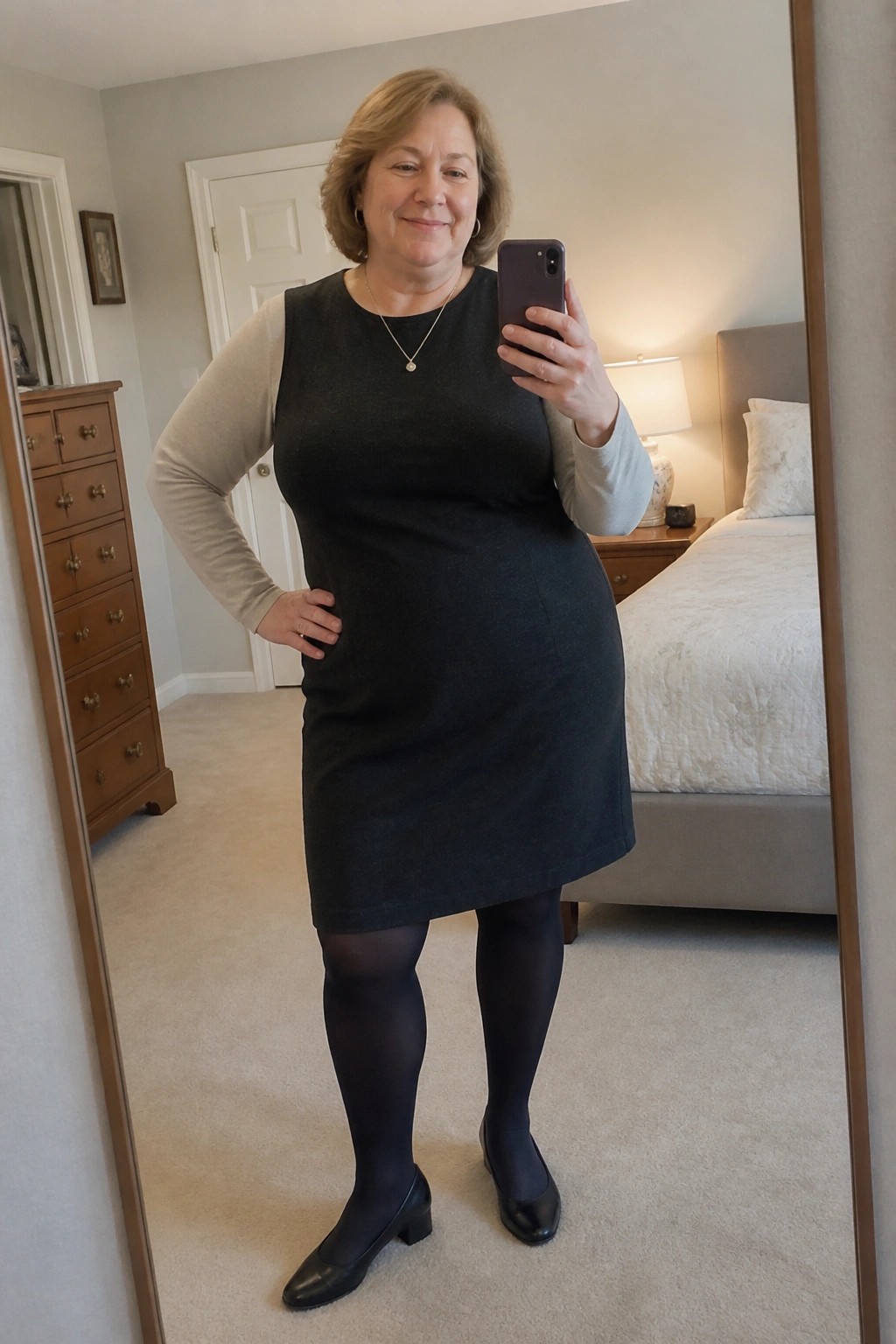 Woman in black dress over beige top taking bedroom mirror selfie