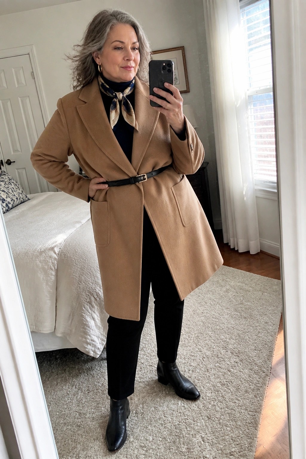 Woman with gray hair in tan coat and black outfit taking mirror selfie in bedroom