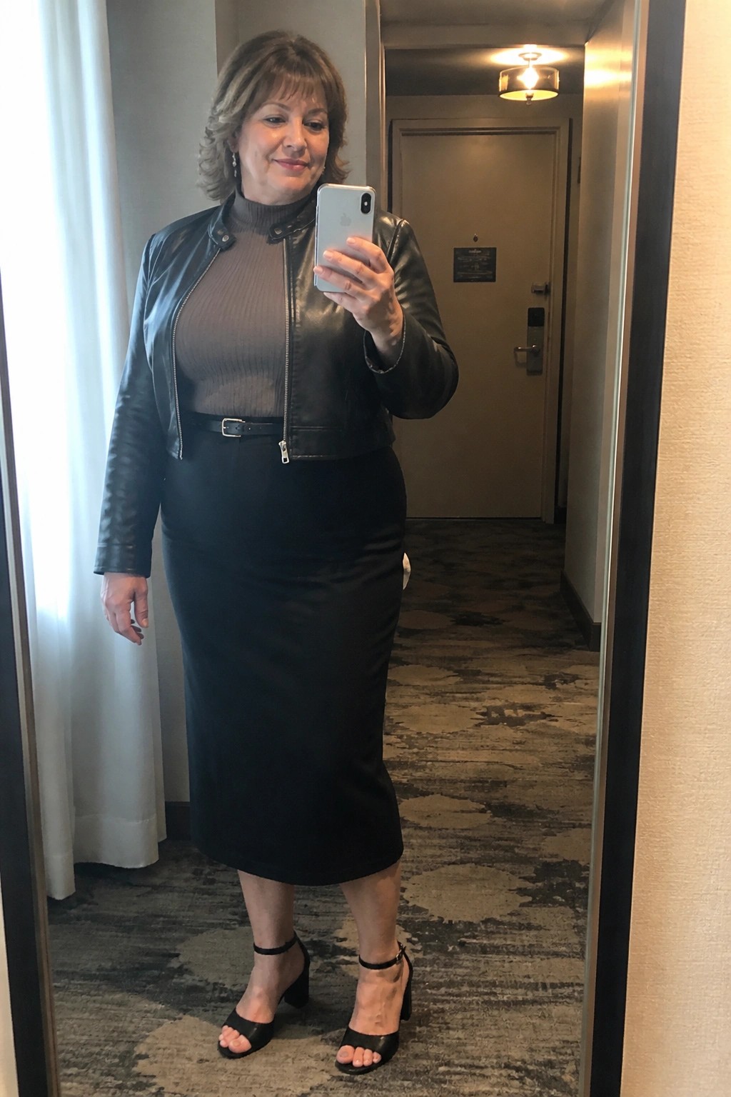 Woman in black leather jacket and skirt taking mirror selfie in hotel hallway