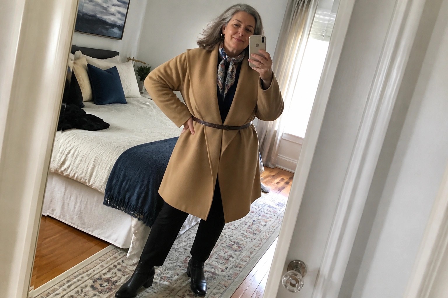 Woman with gray hair in tan coat and black outfit taking mirror selfie in bedroom