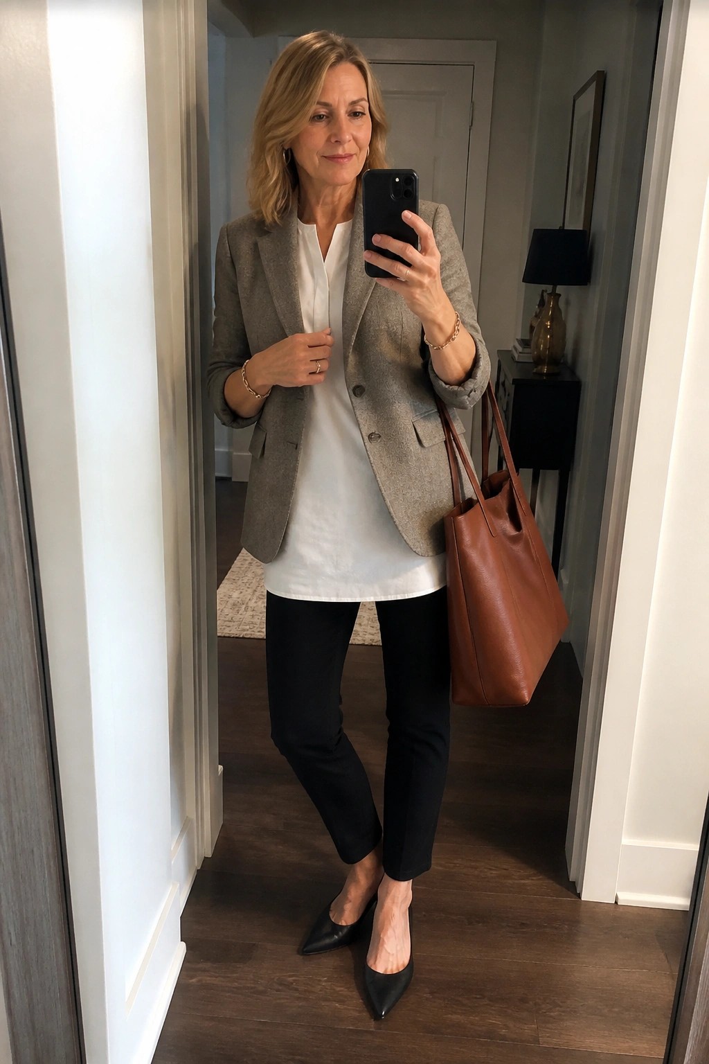 Woman in gray blazer and white top holding phone with brown tote in hallway