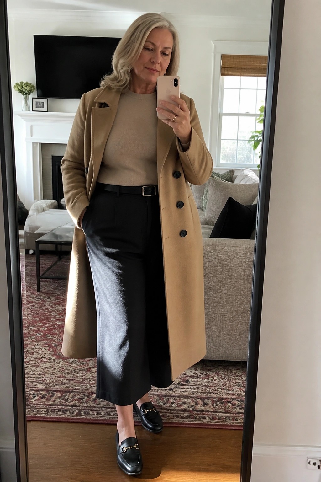 Gray-haired woman in tan coat, beige sweater, and black pants taking mirror selfie.