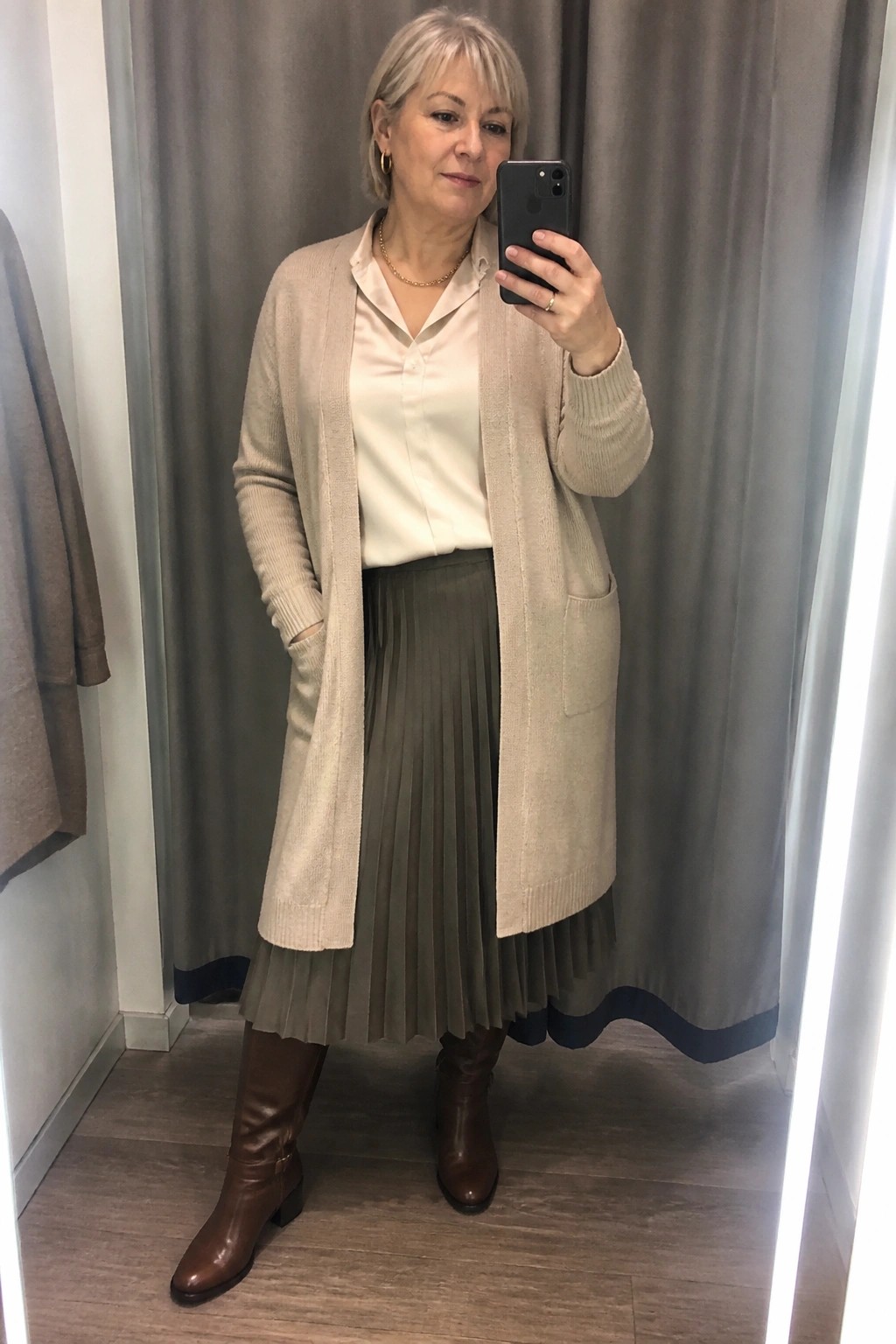 Woman over 50 in plus size outfit wearing long beige cardigan over cream satin shirt and olive pleated midi skirt with brown knee high boots.
