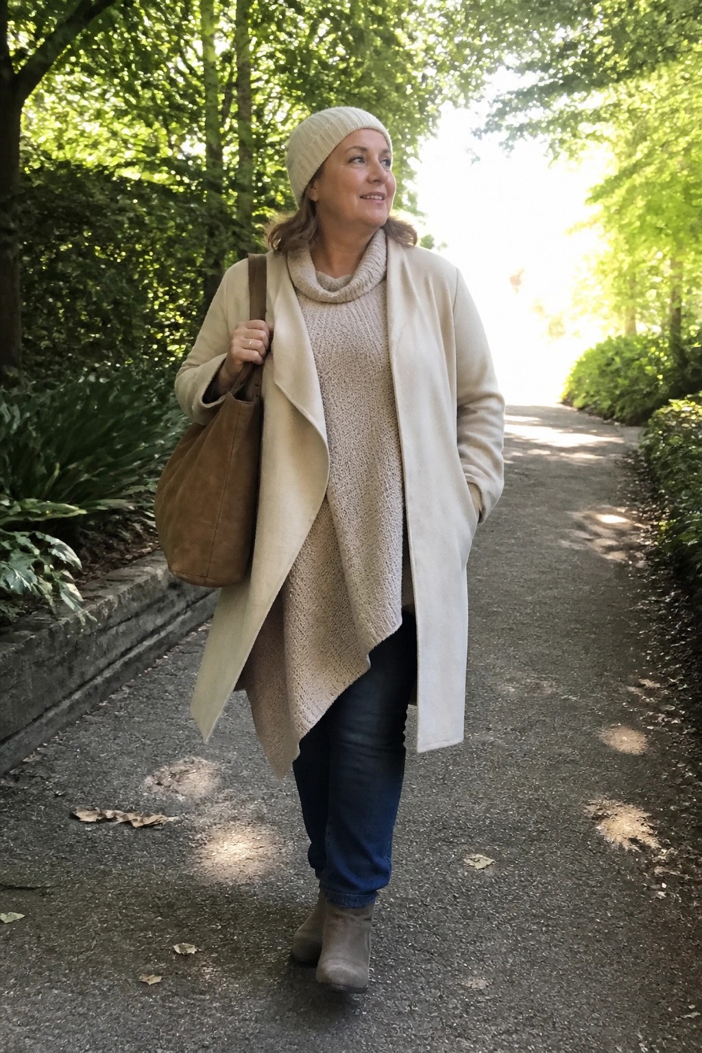 Woman in plus size outfit wearing open long cream coat over beige cowl neck knit sweater, blue jeans, beige suede ankle boots, beige knit beanie, and brown suede shoulder bag on a paved path.