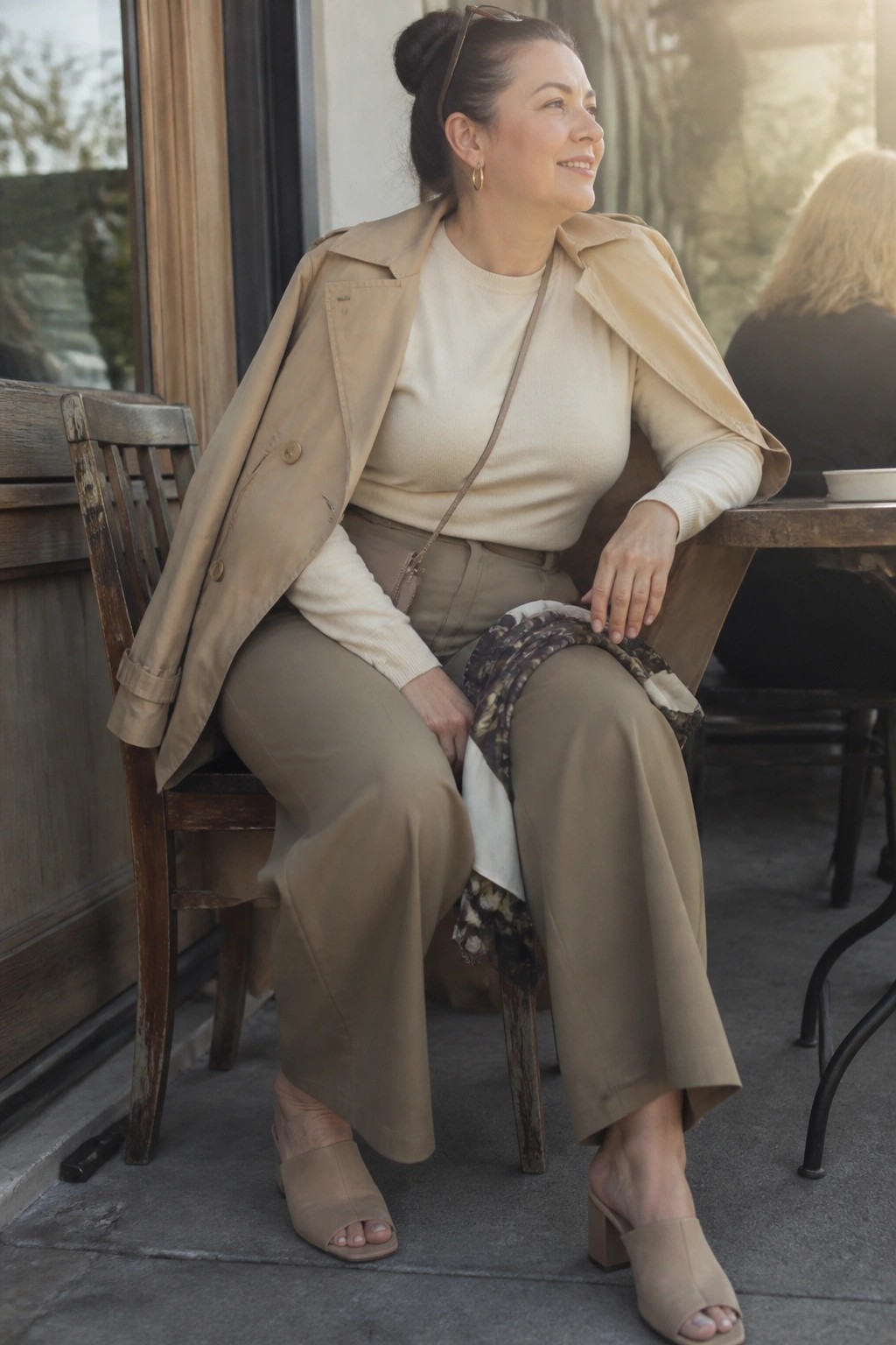 Woman in beige trench coat and trousers seated on wooden chair at outdoor table