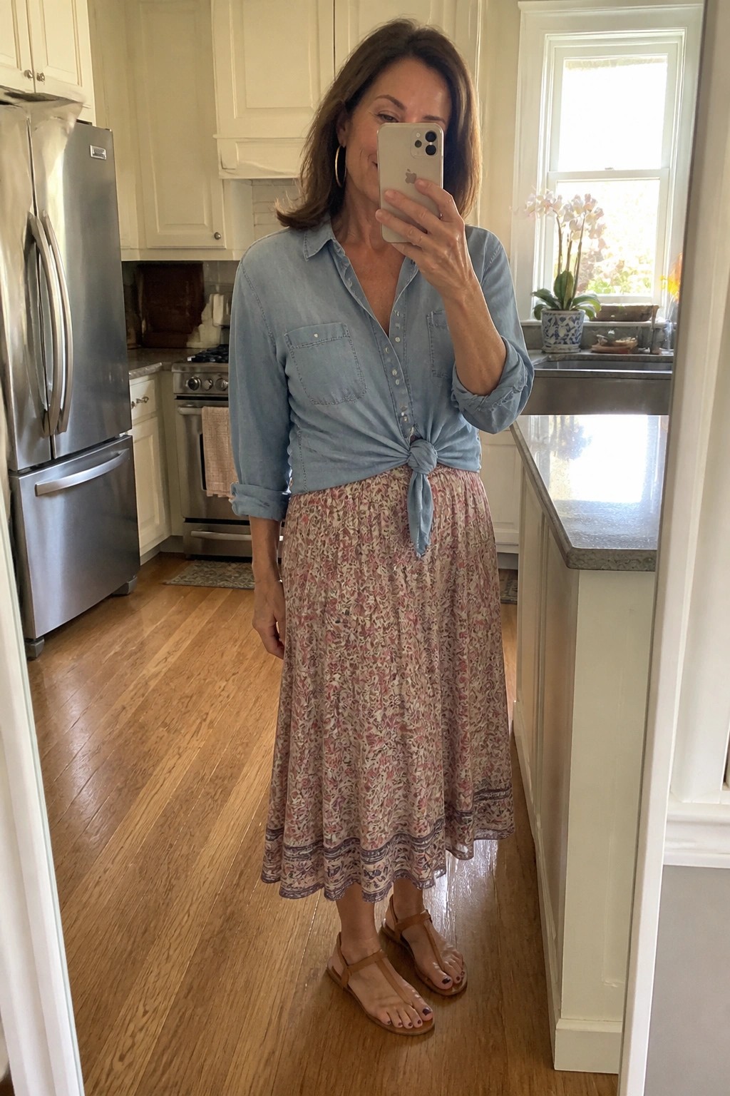 Woman over 50 in light blue chambray shirt knotted at waist over pink floral midi skirt paired with brown strappy sandals standing in kitchen.